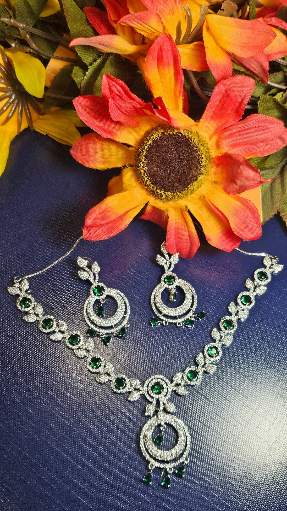 Silver-tone necklace and matching earrings with green accents, leaf-shaped links, open circular pendants and dangling drops, arranged beside decorative flowers.