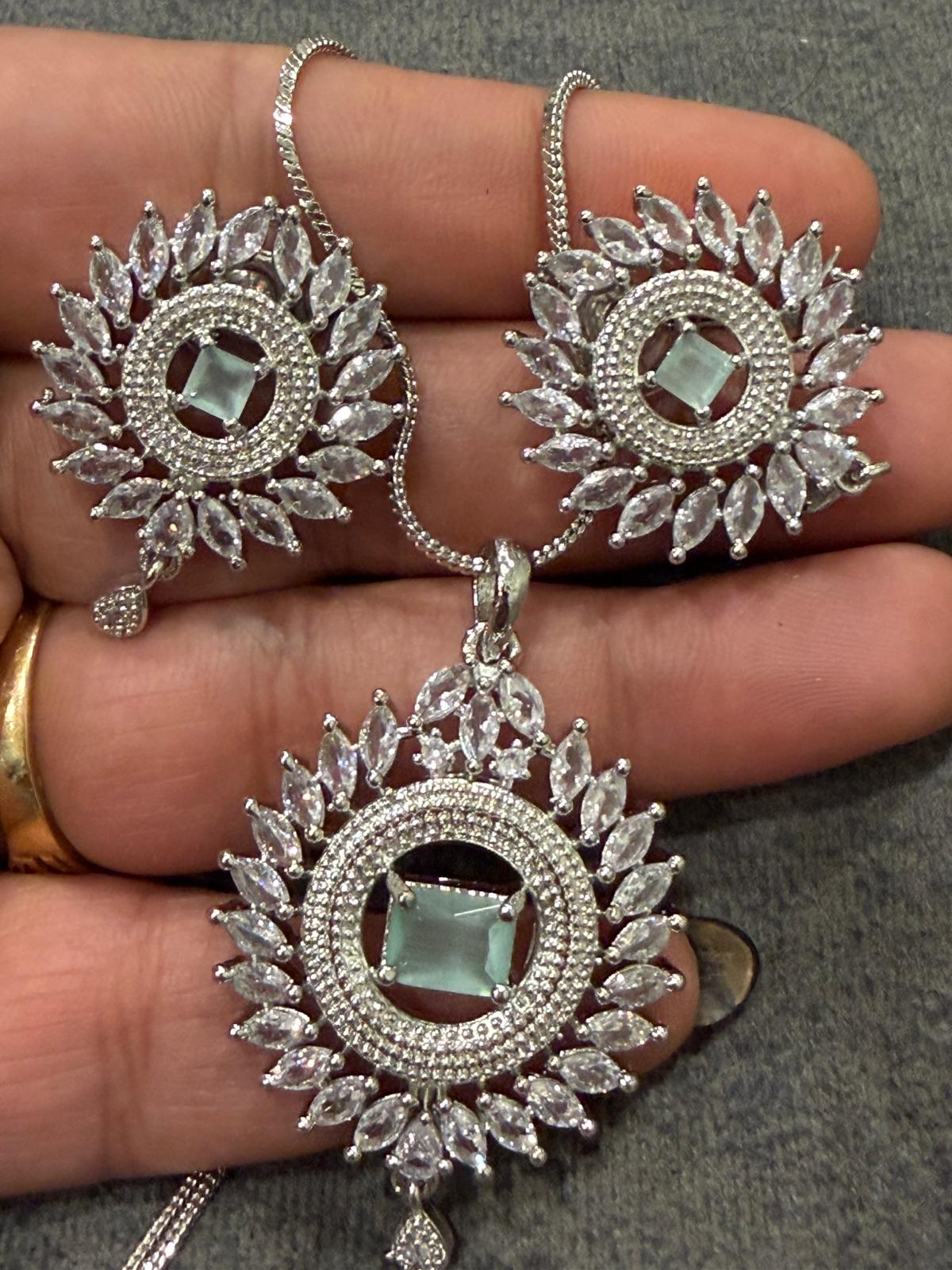 Silver-tone sunburst pendant and two matching earrings with mint green rectangular centers, clear accents and small drop details, shown on a hand.
