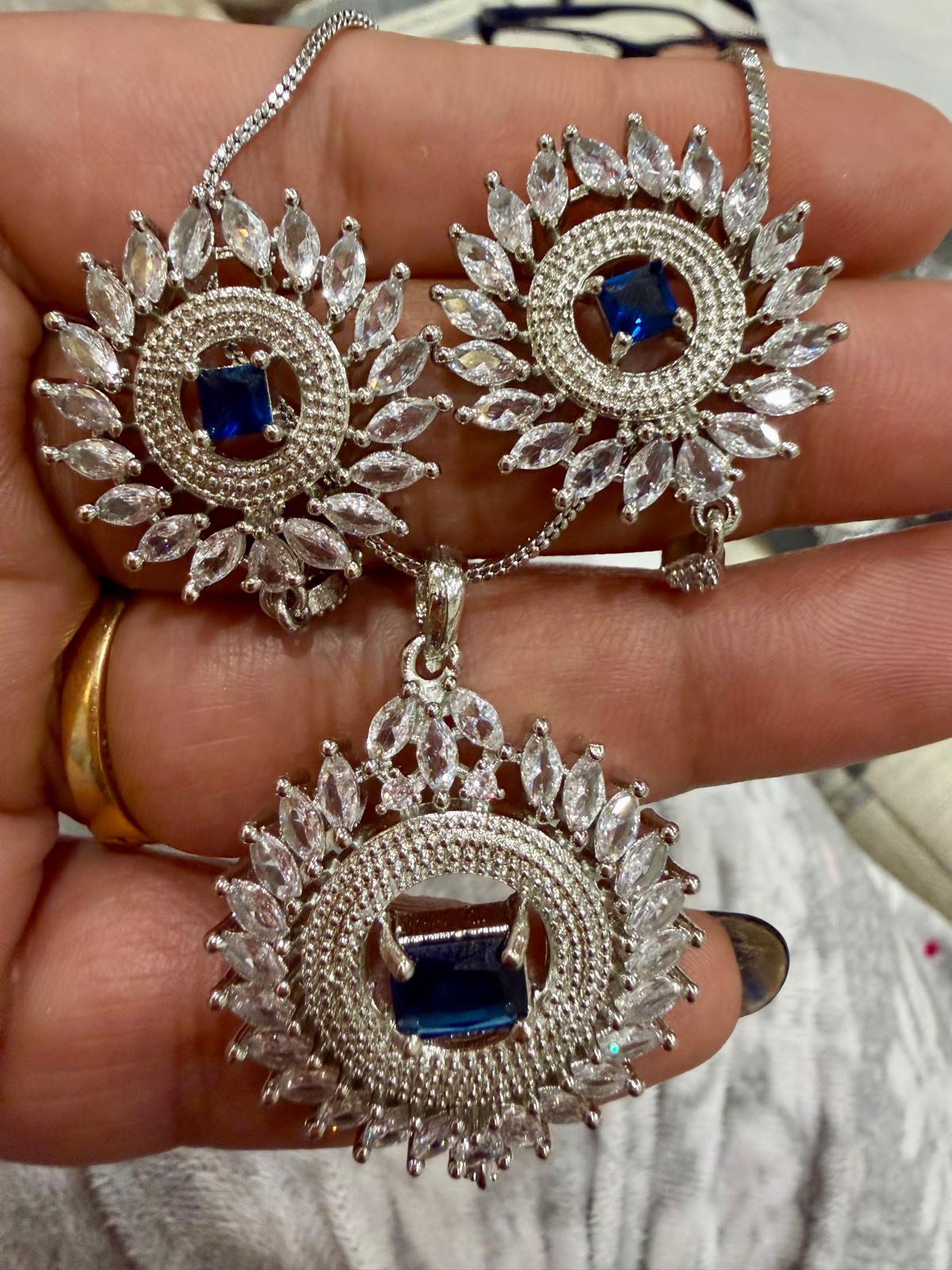 Silver-tone sunburst pendant and two matching earrings with royal blue rectangular centers, clear accents and small drop details, shown on a hand.