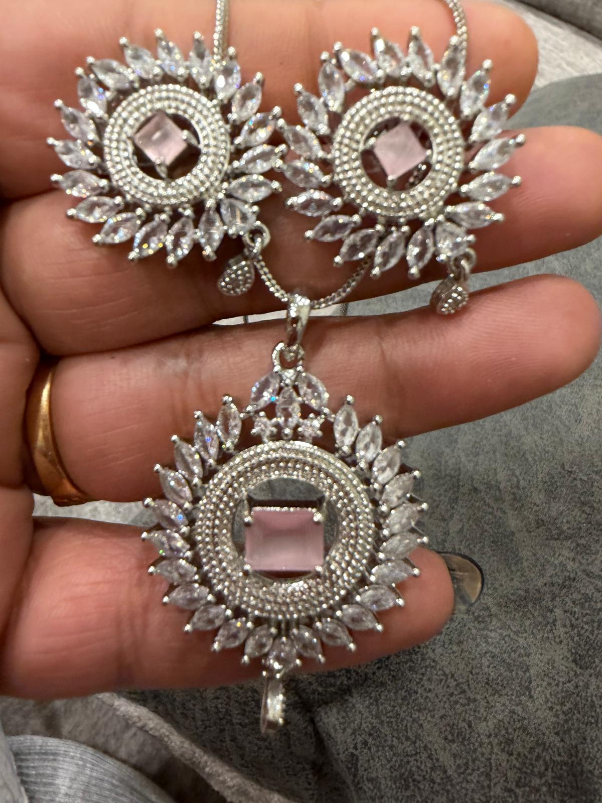 Silver-tone sunburst pendant and two matching earrings with baby pink rectangular centers, clear accents and small drop details, shown on a hand.