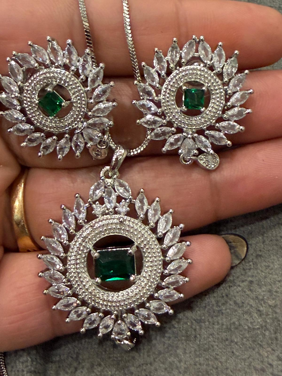 Silver-tone sunburst pendant and two matching earrings with green rectangular centers, clear accents and small drop details, shown on a hand.