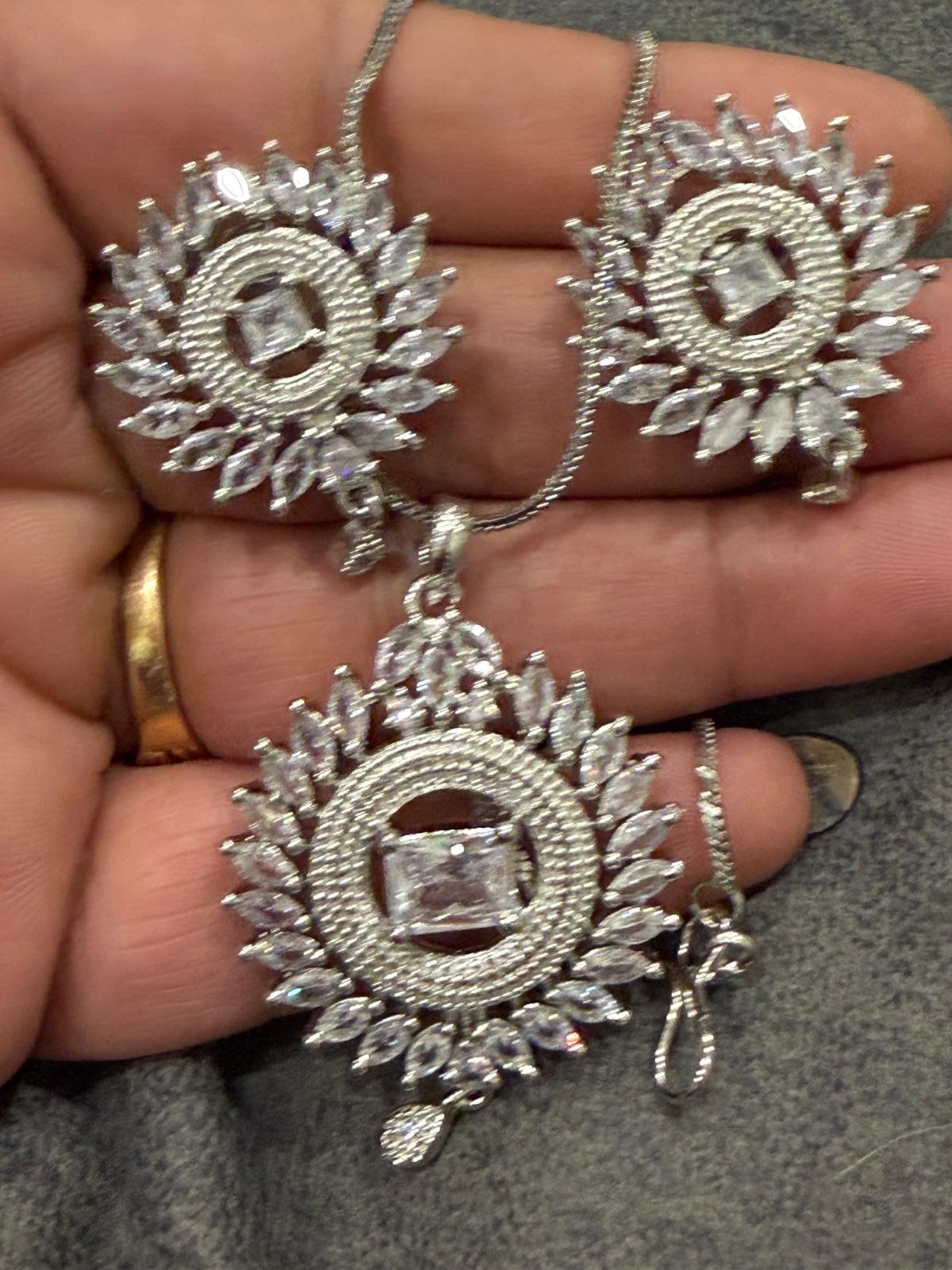 Silver-tone sunburst pendant and two matching earrings with clear rectangular centers, clear accents and small drop details, shown on a hand.