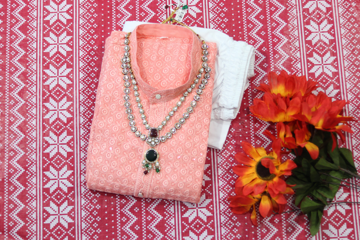 Folded peach kurta with small circular motifs, a band collar and front buttons, arranged beside white fabric and decorative flowers.