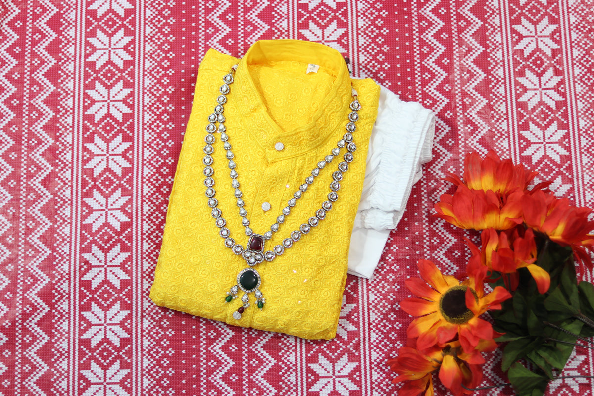 Folded yellow kurta with small circular motifs, a band collar and front buttons, arranged beside white fabric and decorative flowers.