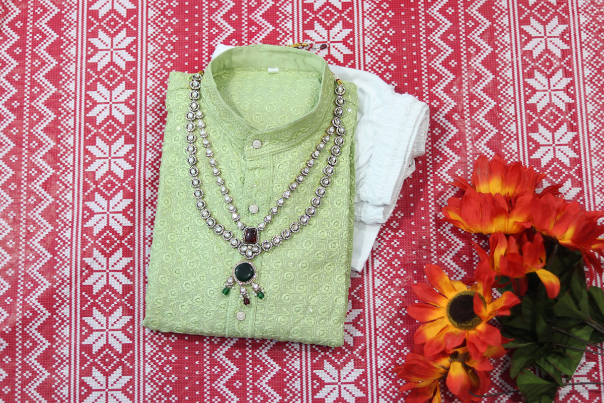 Folded pistachio green kurta with small circular motifs, a band collar and front buttons, arranged beside white fabric and decorative flowers.