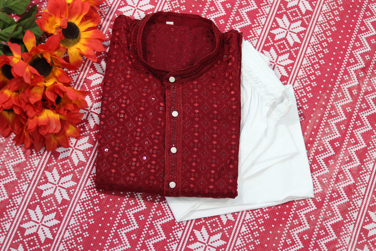 Folded maroon kurta with small geometric motifs, a band collar and front buttons, arranged beside white fabric and decorative flowers.