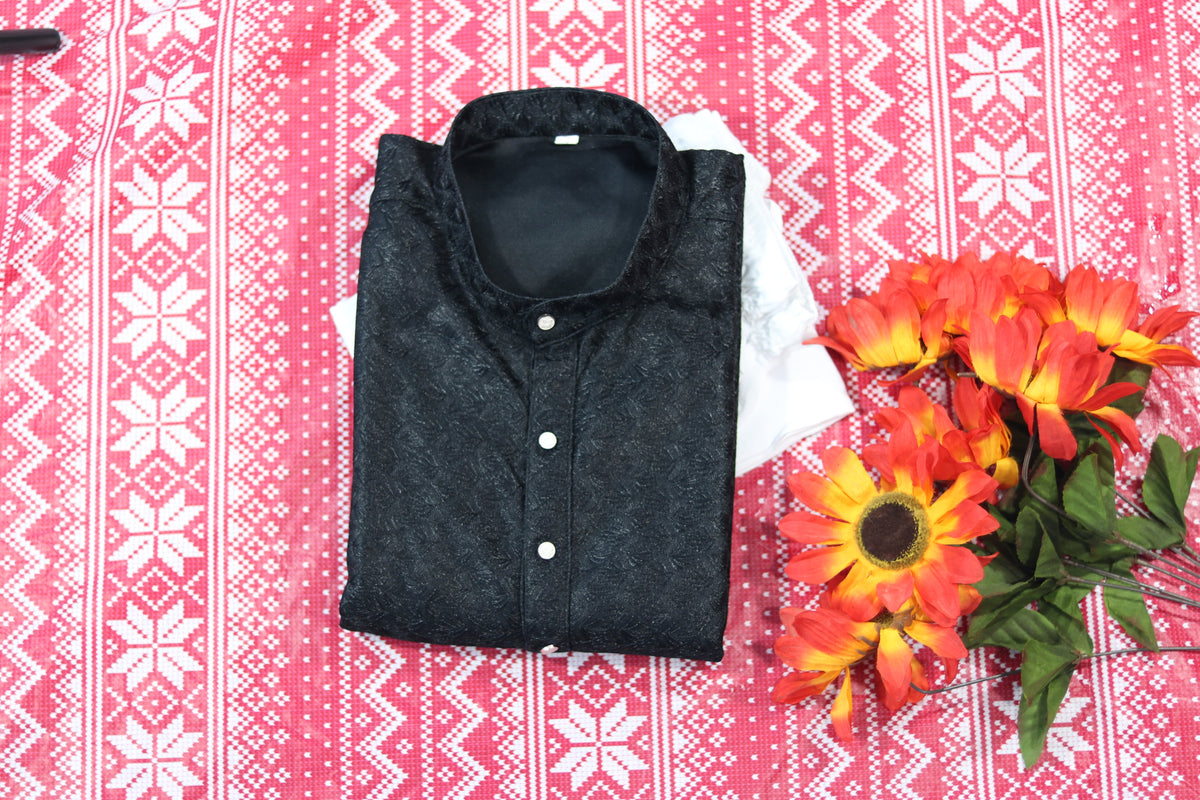 Folded black kurta with fine swirl pattern, a band collar and front buttons, arranged beside white fabric and decorative flowers.