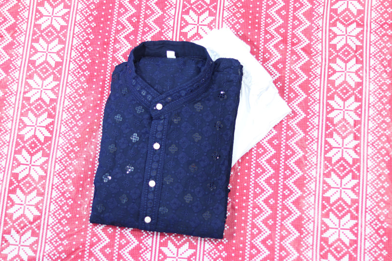 Folded royal blue kurta with floral diamond pattern, a band collar and front buttons, arranged beside white fabric and decorative flowers.